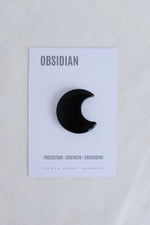 Black crescent moon-shaped obsidian stone on a card with text on a light gray background
